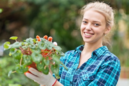 A young woman with blond hair tied back, dressed in a blue and green tartan shirt, is standing outdoors in a garden. She is smiling and holding a potted plant with green leaves and small white flowers. The background features a blurred garden scene with varied foliage, including bushes and trees, under natural daylight with overcast weather. The garden appears well-maintained, with a mix of cultivated plants and natural elements, reflecting typical gardening activities in the outdoor space of a residential property in Chelsea or nearby areas serviced by Landscapers Chelsea. The scene emphasizes the nurturing aspect of gardening and outdoor plant care, suitable for a landscaping or gardening service website focused on garden planting and maintenance in London postcodes.
