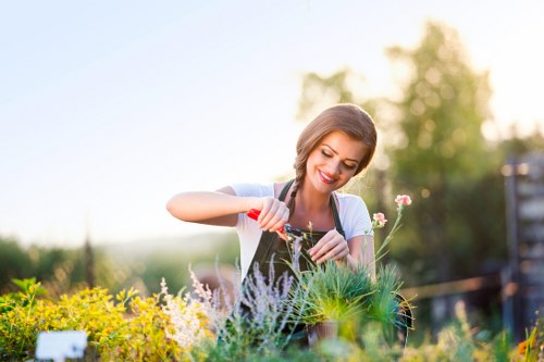 A young woman is tending to a vibrant garden in a back outdoor space near Chelsea, surrounded by various flowering plants, green shrubs, and a grassy lawn area. She is smiling and using watering cans to care for the plants, which include a mix of colorful flowers and lush foliage. The garden features a combination of paved pathways and soil bed borders, with an assortment of textures from the smooth paving stones to the textured plant leaves. In the background, there are trees and a wooden fence, indicating a well-maintained, private outdoor environment. Soft natural sunlight illuminates the scene, suggesting a clear, mild day suitable for gardening activities. This setting demonstrates typical garden features that a landscaping service like Landscapers Chelsea might work on, focusing on outdoor beauty and garden health maintenance in the local area, specifically near the Chelsea postcode district.