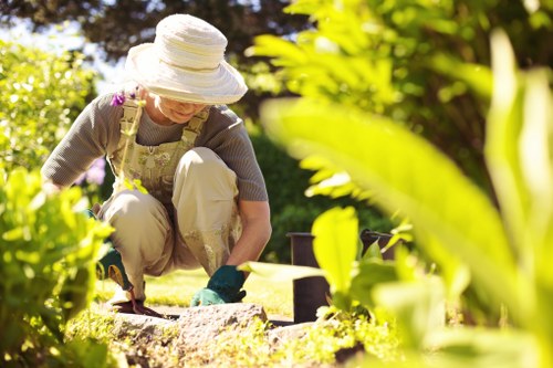 A woman gardening outdoors in a well-maintained backyard with lush green grass and a variety of plants. She is wearing a wide-brimmed sun hat, safety gloves, and casual gardening clothes, and she is kneeling on the lawn while using a trowel to tend to a flower bed bordered by soil and small stones. In the background, there are tall trees and a neatly trimmed hedge, providing natural shade and privacy. The garden features a mix of flowering plants and shrubbery, creating a colourful and inviting outdoor space. Paving stones or wooden decking are partly visible, indicating an area for outdoor seating or pathways. The scene is illuminated by natural sunlight, suggesting a bright, clear day suitable for gardening activities. This image relates to professional landscaping and gardening services offered by Landscapers Chelsea, highlighting expertise in outdoor garden design and maintenance in Surrey, UK.
