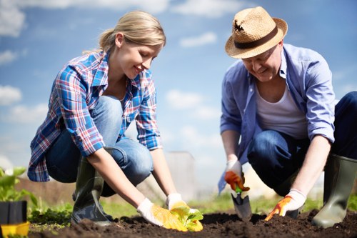A young woman and an older woman are working together in a well-maintained garden, tending to the soil in a cultivated flower bed or vegetable patch. The younger woman, dressed in a blue plaid shirt and jeans, is kneeling on the ground and planting or weeding, demonstrating attentive care. The older woman, wearing a straw hat, a light blue shirt, and dark trousers, is also kneeling and actively involved in gardening work, possibly planting or tending to the plants. The garden features a neatly prepared soil bed with dark, rich earth, bordered by green foliage and low-growing plants. In the background, there is a bright blue sky with scattered clouds, suggesting clear weather ideal for outdoor gardening. The surroundings imply a well-kept outdoor space, likely part of a residential or community garden in Chelsea, with a natural and vibrant atmosphere. The scene reflects the typical activities of planting and garden maintenance that landscaping services like those offered by Landscapers Chelsea might support, highlighting a connection to local outdoor gardening expertise.
