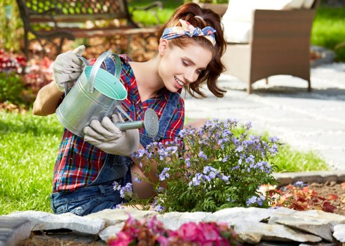 A young woman with a joyful expression is gardening outdoors in a residential garden, watering a small flowering shrub with a light green metal watering can. She is dressed casually in a colorful plaid shirt, denim shorts, and grey gardening gloves, with a floral headband in her brown hair. The garden features a neatly maintained lawn in the foreground, bordered by a low stone edge that separates it from flower beds filled with vibrant pink and purple perennial plants. In the background, there is a wooden outdoor table and chairs, a paved patio area, and mature trees providing shade. The scene suggests a bright, sunny day with natural light illuminating the lush greenery and colorful blooms, reflecting a well-kept outdoor space suitable for gardening and outdoor relaxation, as offered by landscaping services from Landscapers Chelsea in the London SW10 area.