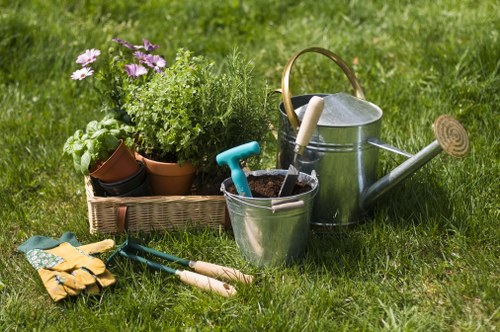 A neatly maintained garden scene featuring a lush green lawn bordered by a variety of potted plants and flower beds, including purple, green, and white foliage plants. The foreground shows gardening tools such as a pair of garden gloves, small hand trowels, and pruning shears resting on the grass. On the right side, a classic metal watering can with a wooden handle and a long spout is positioned near a galvanized bucket containing soil and a small gardening trowel. In the background, there is a low hedge providing a natural boundary and a backdrop of tall grass. The lighting suggests a bright, fair-weather day with natural sunlight illuminating the vibrant greenery and gardening equipment, creating an inviting outdoor environment suitable for garden maintenance and landscaping projects. Gardens like this are typical in residential outdoor spaces within Chelsea and surrounding areas, where professional gardening services by Landscapers Chelsea can enhance outdoor aesthetics and promote healthy plant growth.