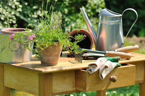 A wooden garden table situated outdoors in a lush green garden features various gardening tools and potted plants. On the table, there are terracotta and ceramic pots containing green, leafy plants, with some flowering varieties showing small pink blooms. A metal watering can with a curved handle and spout is positioned towards the right side of the table, alongside a small garden rake. A pair of gardening gloves, slightly rumpled, rests atop the table near the edge, indicating recent use. The surface of the table displays a natural, weathered wood grain, and underneath is a background of dense foliage, including trees and shrubs, suggesting a well-maintained backyard garden in a rural or suburban area. The overall scene highlights a calm, sunny day with natural light illuminating the tools and plants, reflecting a typical gardening activity setting in Chelsea or nearby areas, supporting professional gardening and landscaping services offered by Landscapers Chelsea. The environment emphasizes a tidy and thriving outdoor space suited for detailed garden maintenance and planting tasks.