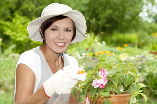 A woman with short brown hair, wearing a wide-brimmed white gardening hat, white gloves, and a light grey t-shirt, is tending to a potted plant with pink flowers in a lush garden setting. The garden features a well-maintained lawn with dense green grass in the foreground, bordered by flower beds and natural soil patches. Behind her, there are a variety of mature trees and shrubs, creating a shaded, vibrant outdoor environment typical of landscaped gardens in Chelsea. The scene is lit by natural daylight under a slightly overcast sky, highlighting the fresh, natural tones of the plants and garden surfaces. This image captures the landscaping and gardening activities associated with maintaining attractive outdoor spaces, relevant to professional gardening services offered by Landscapers Chelsea in the local area, including Chelsea and the surrounding postcode area.