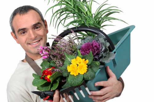 The image shows a man with short dark hair smiling while holding a teal plastic garden trowel and supporting a round black container filled with various flowering plants and greenery. The plants include purple and pink blooms, yellow flowers, and lush green leaves, suggesting a vibrant and well-maintained garden arrangement. Behind him, a large potted plant with long, arching green leaves is visible, placed on a paved outdoor surface area. The background appears to be a garden or outdoor landscape, with the scene suggesting active gardening or plant care activities typical of professional landscaping services in Chelsea or nearby areas. The natural lighting indicates a bright day, ideal for outdoor gardening tasks. This image illustrates gardening tools and plant care, aligning with services offered by companies like Landscapers Chelsea specializing in garden design and maintenance.