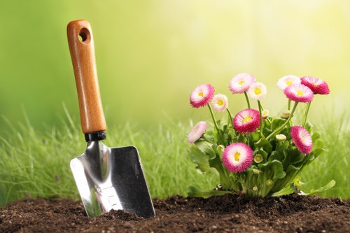 A close-up of a gardening scene in a backyard or front garden, featuring a small garden bed with dark, moist soil and a collection of pink and white flowers with rounded petals, possibly self-seeding annuals or bedding plants, growing densely with green foliage. To the left, a small hand trowel with a wooden handle and metal blade is partially inserted into the soil, indicating recent planting or soil tending. In the background, a lush, well-maintained lawn with vibrant green grass extends towards a softly blurred backdrop of trees or hedges, suggesting a tidy, landscaped outdoor space in a residential setting potentially within Chelsea or nearby postcode areas, managed by Landscapers Chelsea. The scene is evenly lit with natural daylight, suggesting mild weather and no harsh shadows, emphasizing the healthy condition of the plants and the readiness for further gardening or lawn care activities. This image typically represents a well-kept garden during a planting or maintenance session, aligning with their gardening and landscaping services in London.