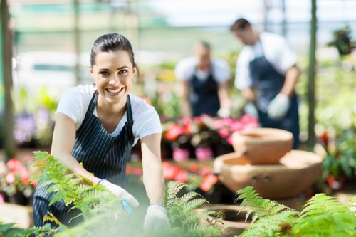 A smiling woman wearing a white t-shirt and striped gardening apron is working in a greenhouse or nursery, surrounded by a variety of potted flowering plants and lush greenery. She is leaning forward with both hands reaching towards the plants, indicating active gardening or planting. In the background, two individuals, also dressed in gardening attire, are engaged in plant care amidst shelves filled with flowers, shrubs, and gardening tools. The environment is bright and well-lit, suggesting daylight filtering through the greenhouse glass, with natural tones of green, pink, and earthy browns dominating the scene. The scene reflects a professional gardening or landscaping setting, emphasizing plant care and outdoor space enhancement, which aligns with services offered by Landscapers Chelsea in the local area around Chelsea, London, or nearby districts. The overall atmosphere is vibrant and professional, suitable for a gardening service website focused on plant nursery care, planting projects, or landscape preparation.